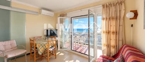 A living room with a sofa, a dining table with chairs, a TV, and a sliding glass door leading to a balcony with a view of the sea.