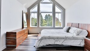 Une chambre avec un plafond en pente, une grande fenêtre donnant sur les arbres, un lit recouvert d’une couvre-lit à motifs et une commode en bois.