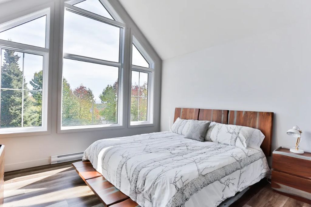 A bedroom with a large window, a bed with a patterned bedspread, a wooden headboard, and a bedside table with a lamp.