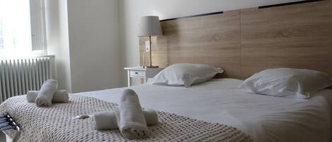 Habitación de hotel con una cama grande, dos almohadas, una mesita de noche con lámpara y una ventana con persianas.