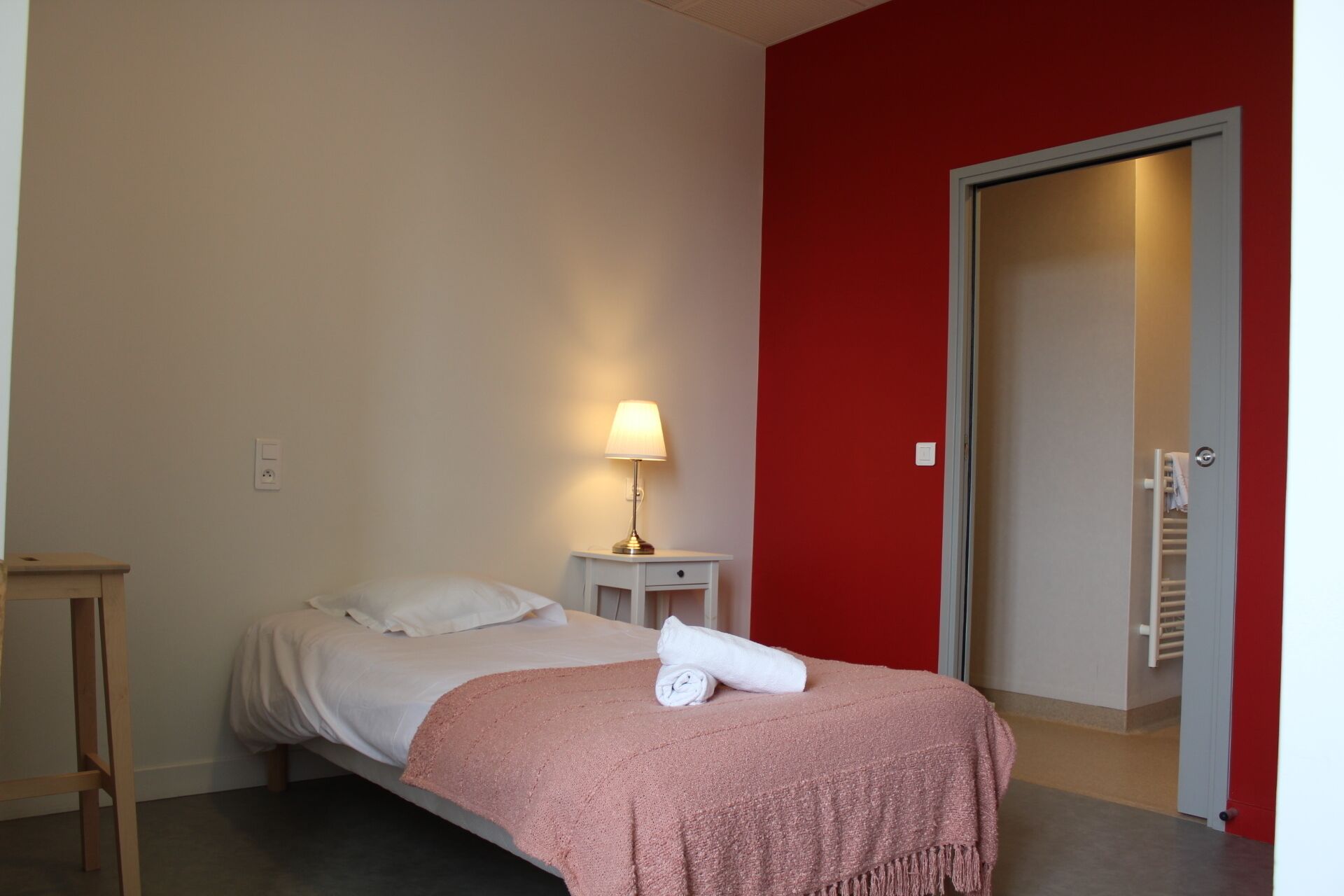 A bedroom with a bed, a bedside table with a lamp, a red wall, and a door leading to a bathroom.