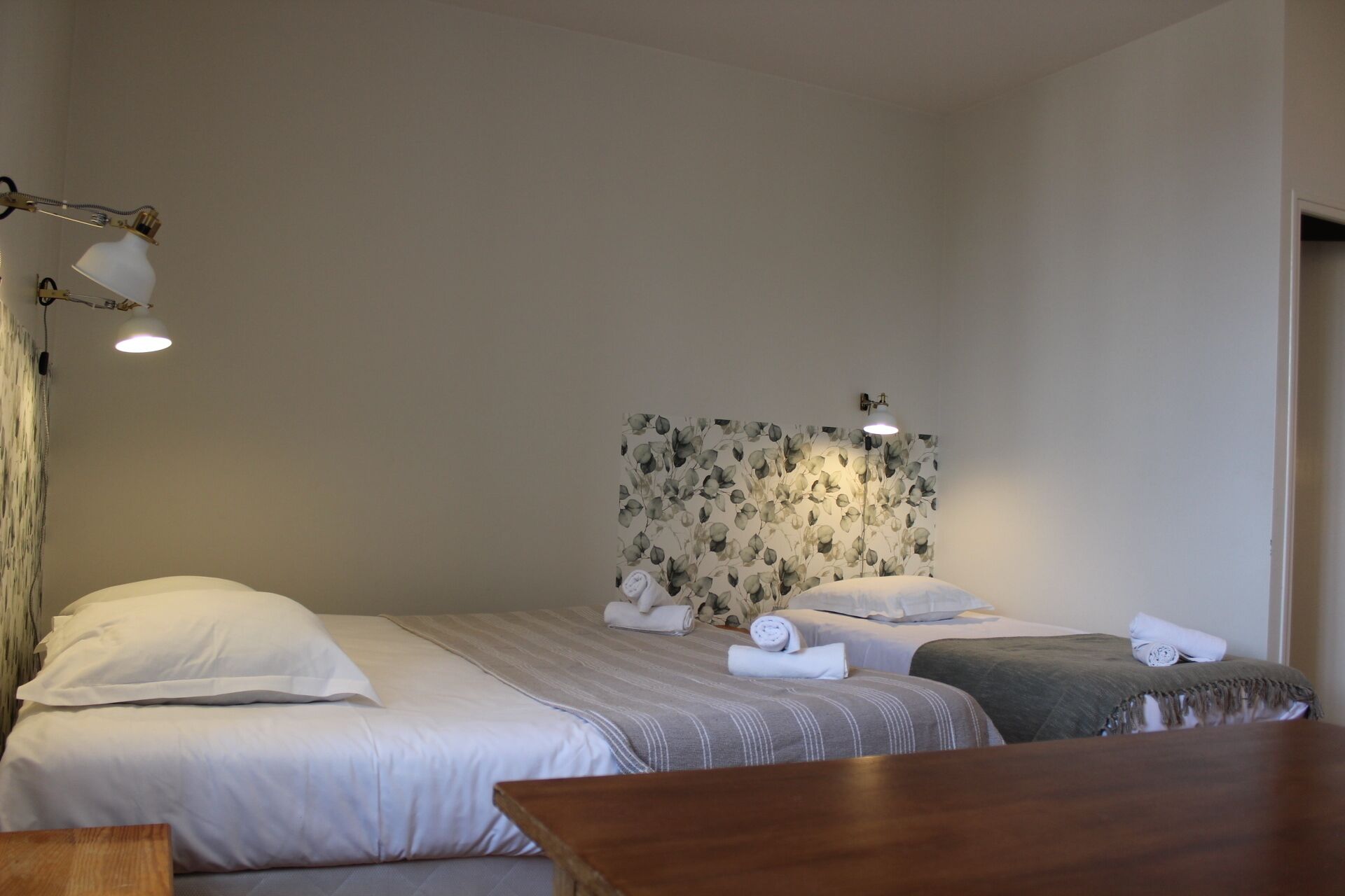 A hotel room with a bed, a bedside table, a headboard with a floral pattern, and a wall-mounted lamp.