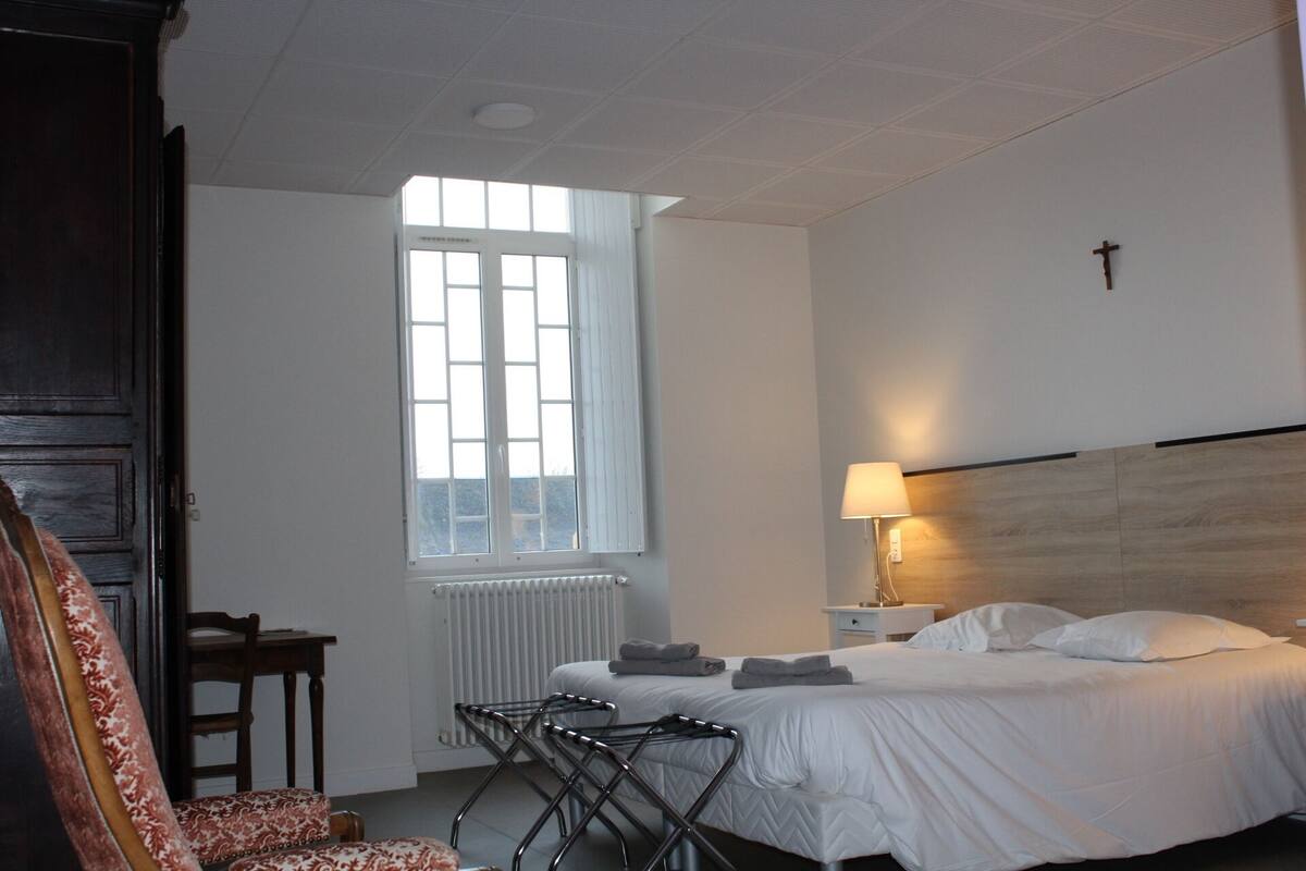 Une chambre d’hôtel avec un lit, un bureau, une chaise, un radiateur et une fenêtre.
