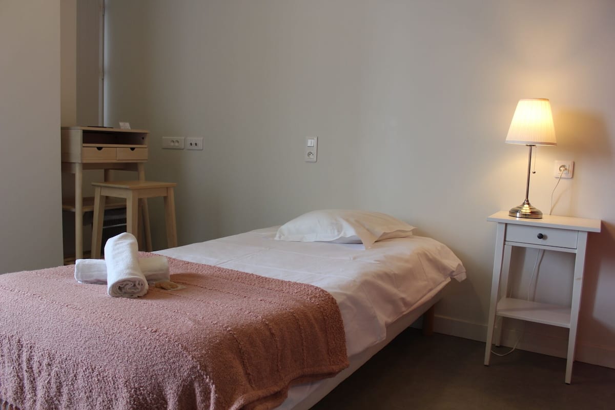 Un lit bien fait, avec une couverture rose, un oreiller blanc et une serviette blanche roulée posée sur le côté. On y trouve une table de chevet avec une lampe et une petite étagère.