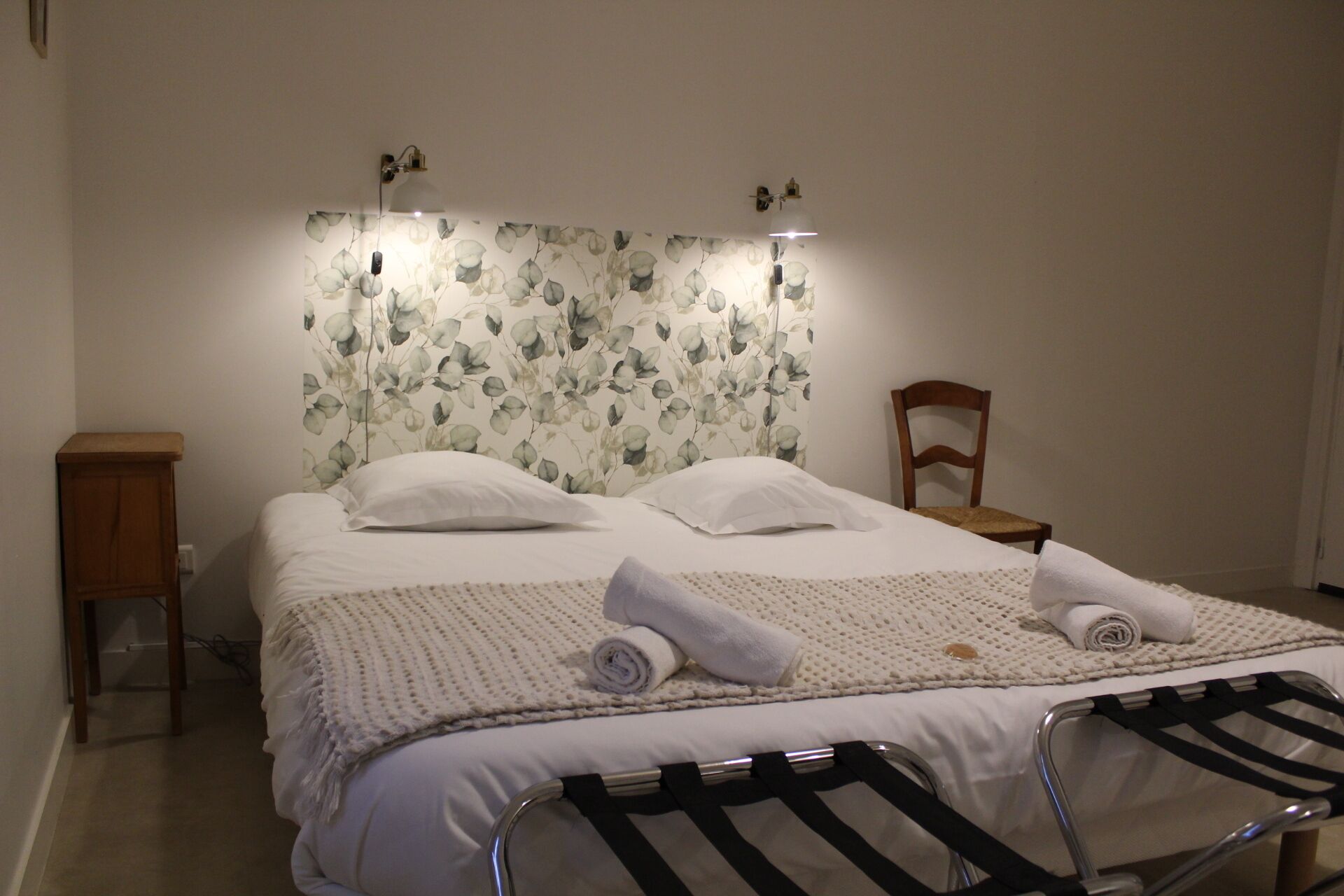 A neatly made bed with white linens and a floral-patterned headboard. There is a wooden nightstand to the left and a chair to the right.