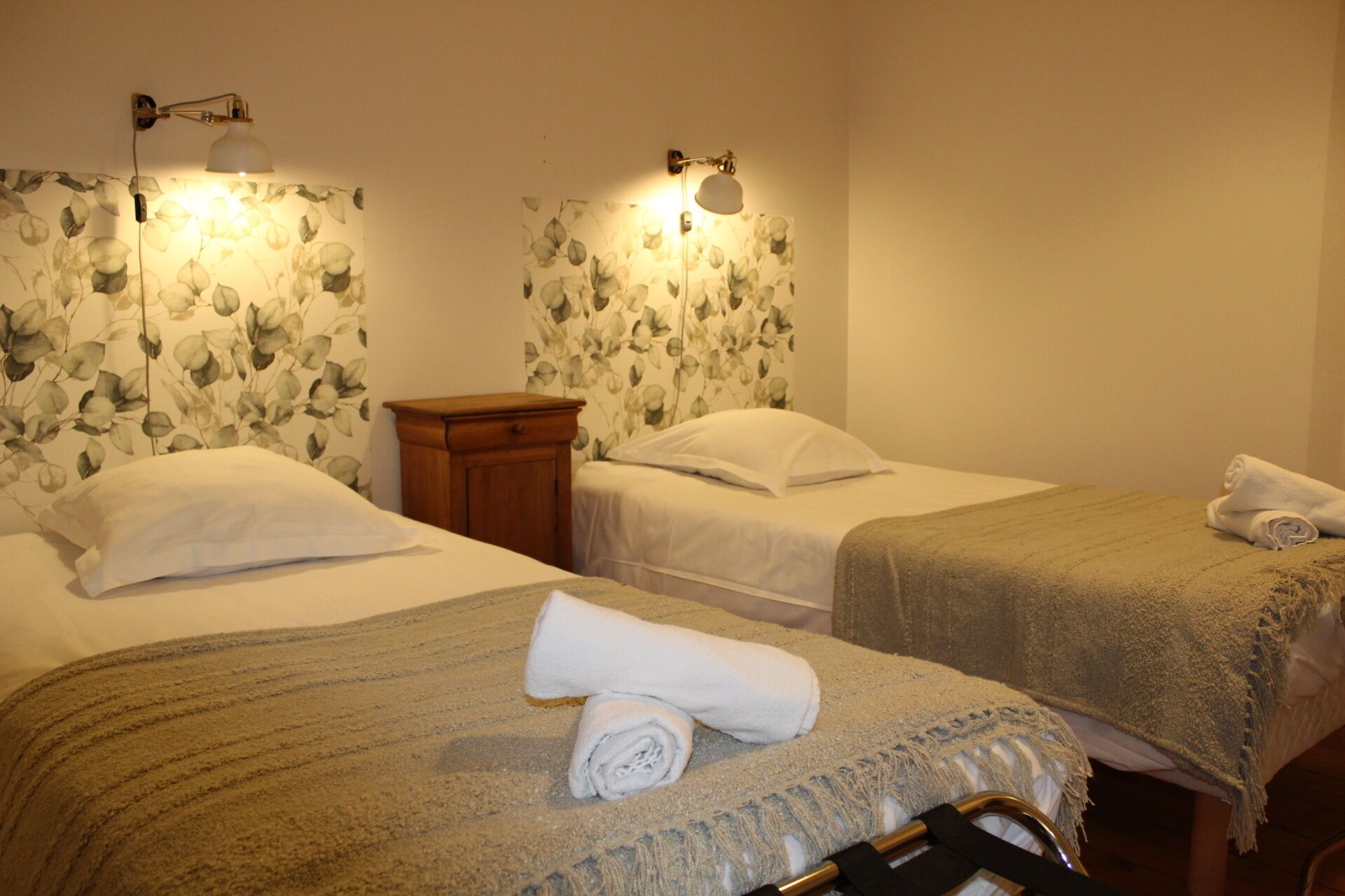 A hotel room with two beds, each with a white pillow and a rolled towel. There are wall-mounted lamps with floral wallpaper behind the beds.