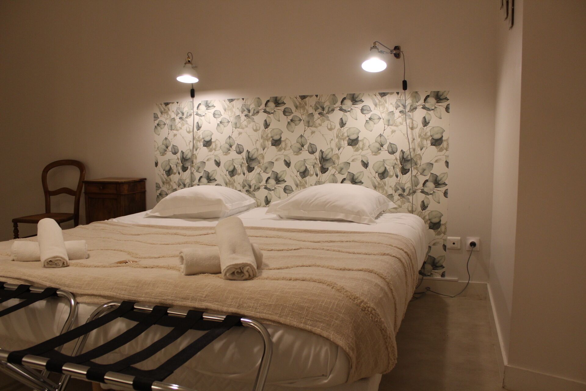 A neatly made bed with white linens and a beige blanket, a wooden chair, and a small bedside table.