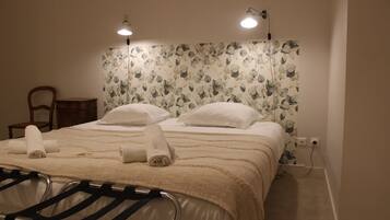 A neatly made bed with white linens and a beige blanket, a wooden chair, and a small bedside table.