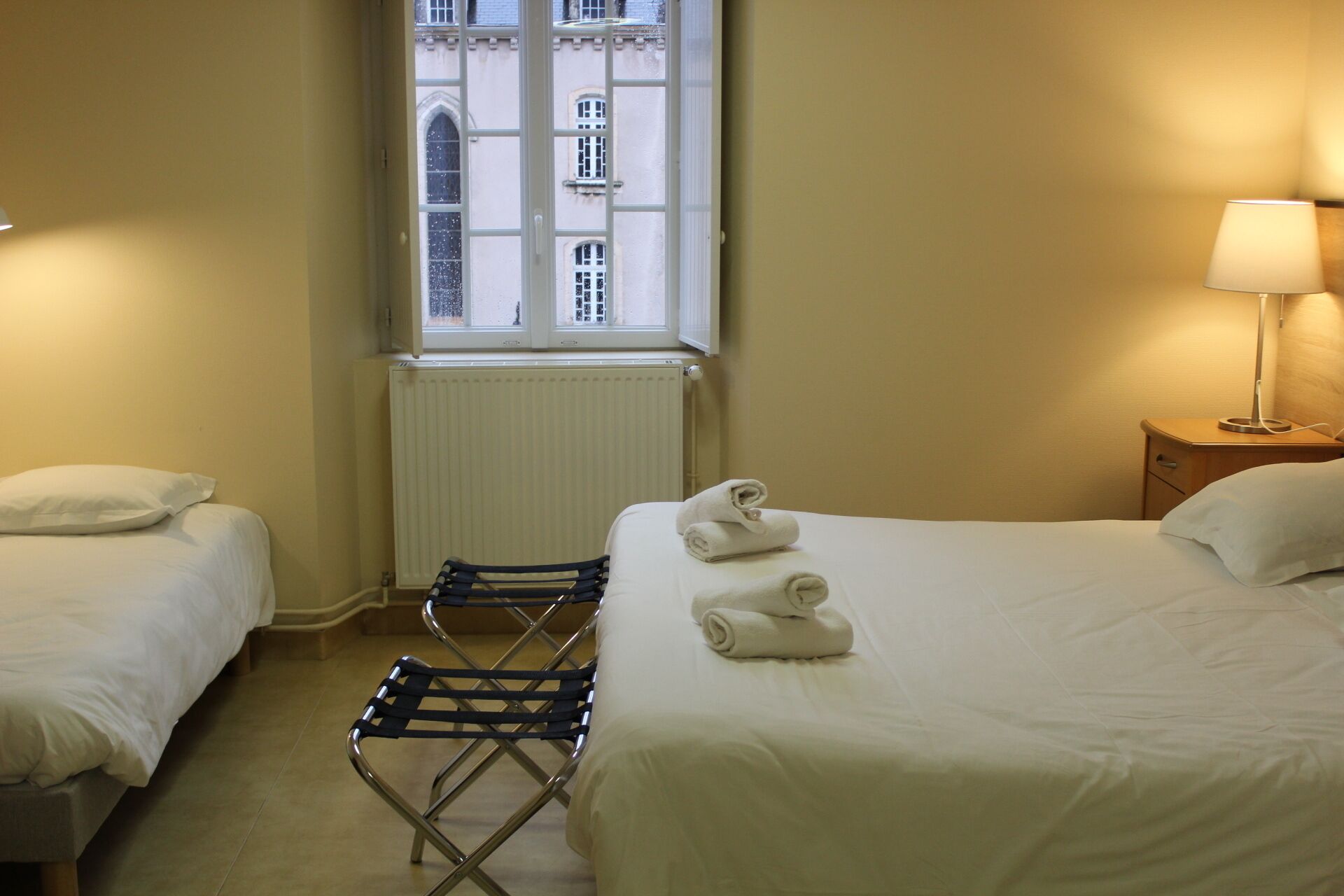 A hotel room with two beds, a radiator, a window with a view of buildings, and a lamp on a bedside table.
