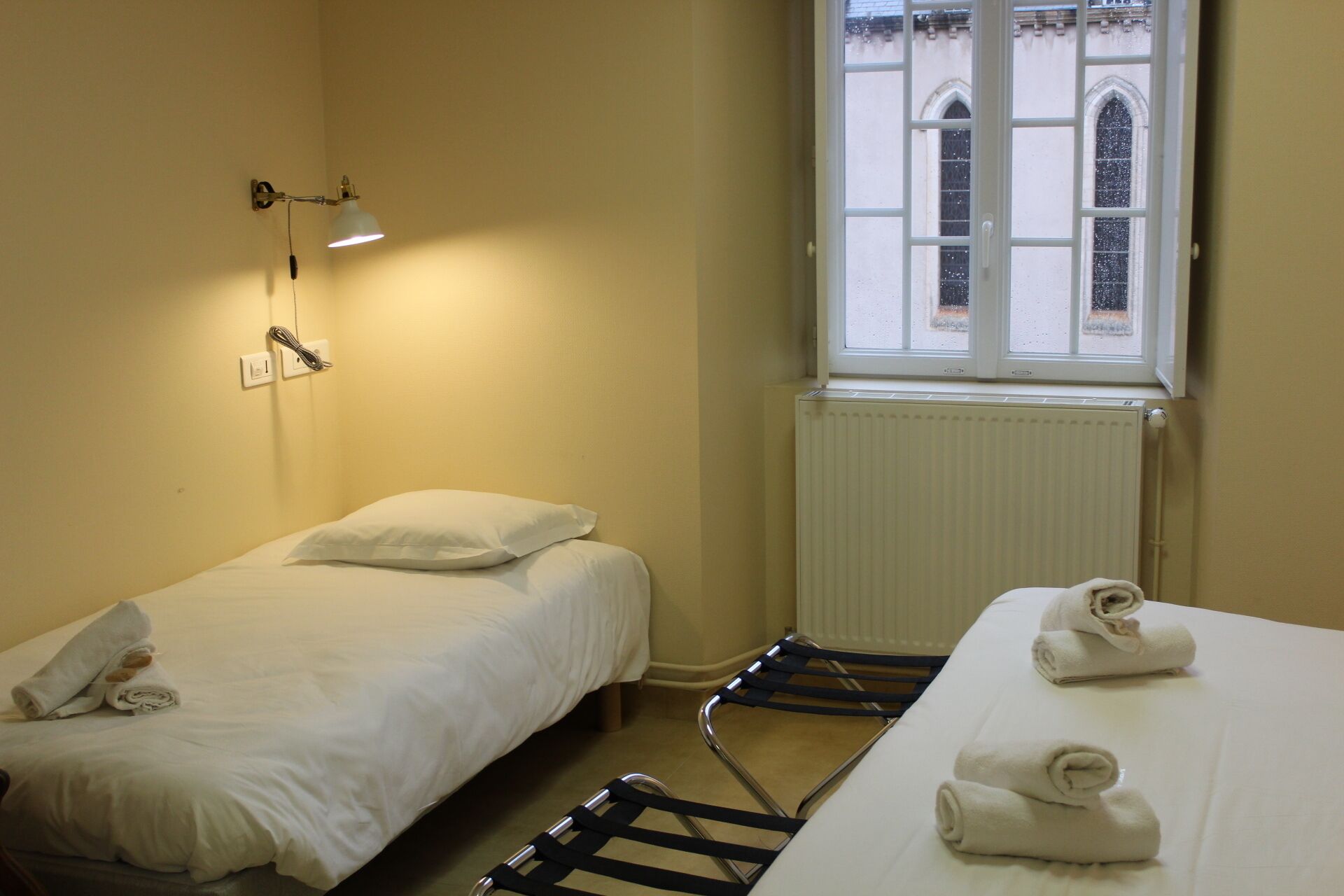A hotel room with two beds, a window with a view of a building, a radiator, and a wall-mounted lamp.