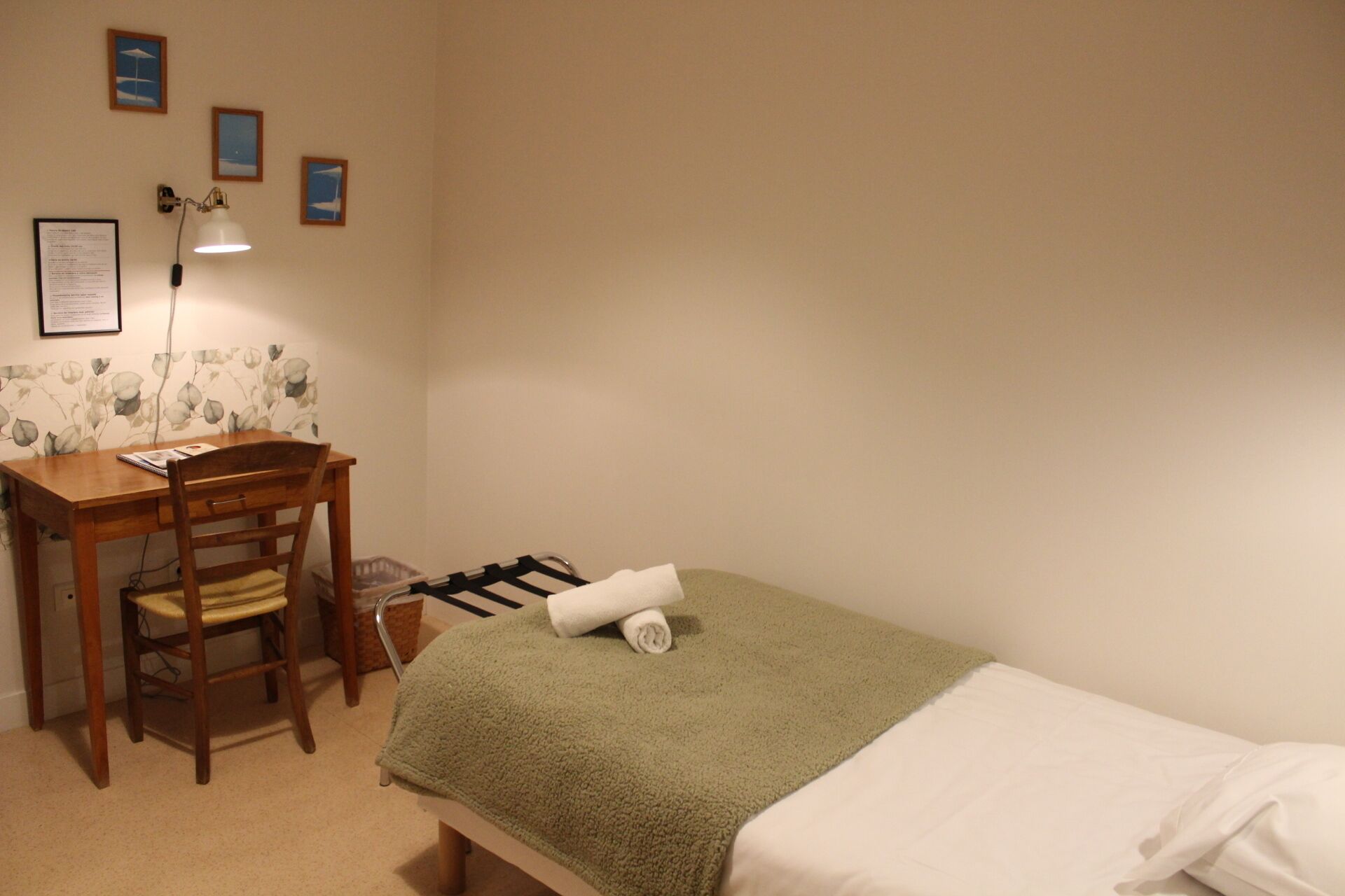 A small room with a bed, a desk, and a chair. There are framed pictures on the wall.