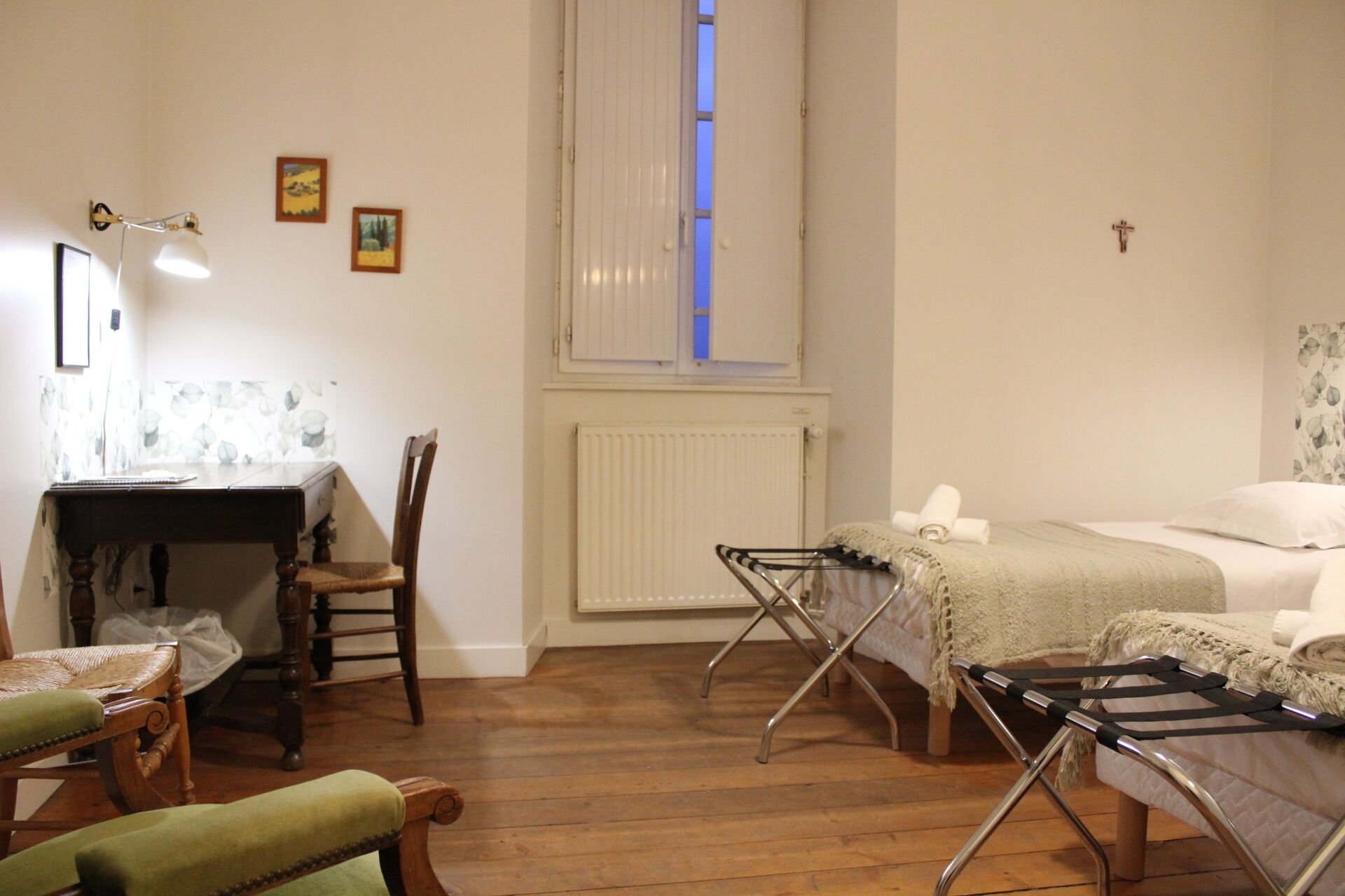 A small, cozy room with a wooden floor, a desk with a chair, a bed with a blanket, and a radiator.