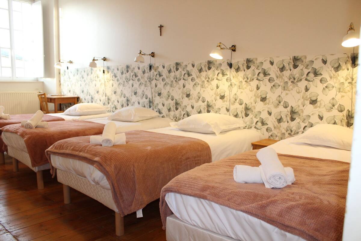 Une chambre d’hôtel avec trois lits, chacun recouvert d’une couverture marron et agrémenté d’oreillers blancs, et un papier peint à motifs floraux sur le mur.