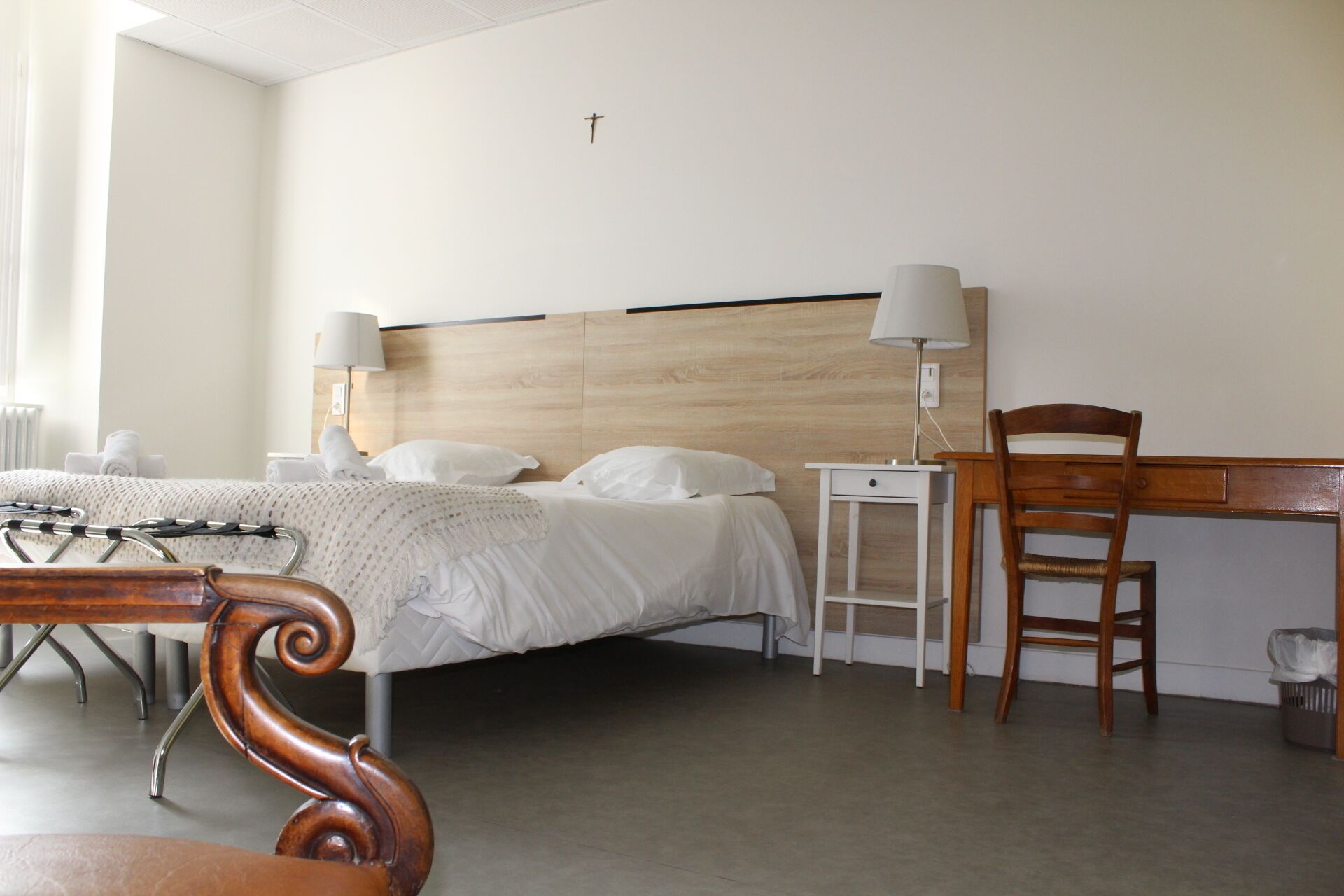 A hotel room with a bed, bedside lamps, a wooden chair, a desk, and a cross on the wall.