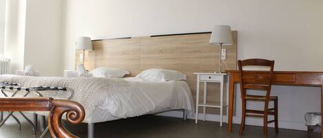 Habitación de hotel con cama, lámparas de noche, una silla de madera, escritorio y una cruz en la pared.