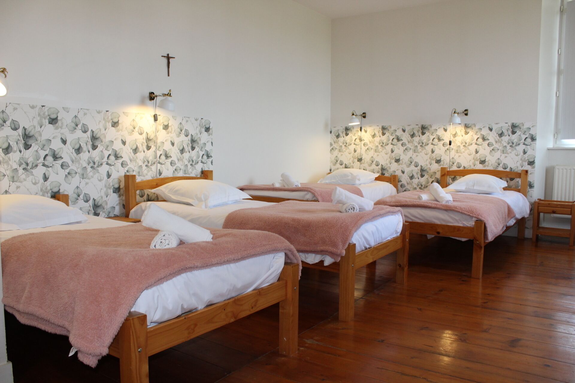 A room with four wooden beds, each with white linens and pink towels. There are wall-mounted light fixtures and a cross on the wall.