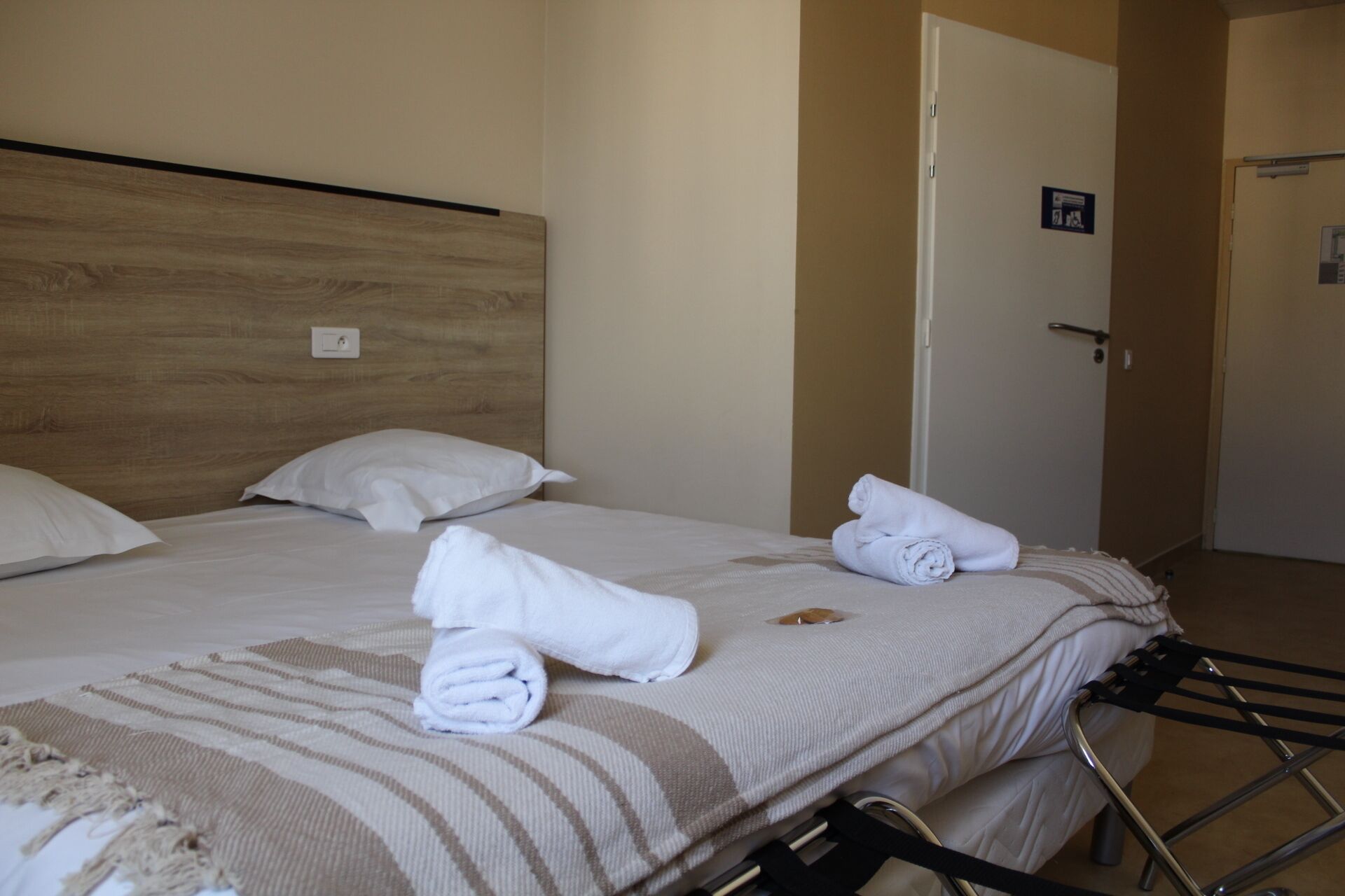 A hotel room with a wooden headboard, two white pillows, a striped blanket, and rolled towels on the bed.