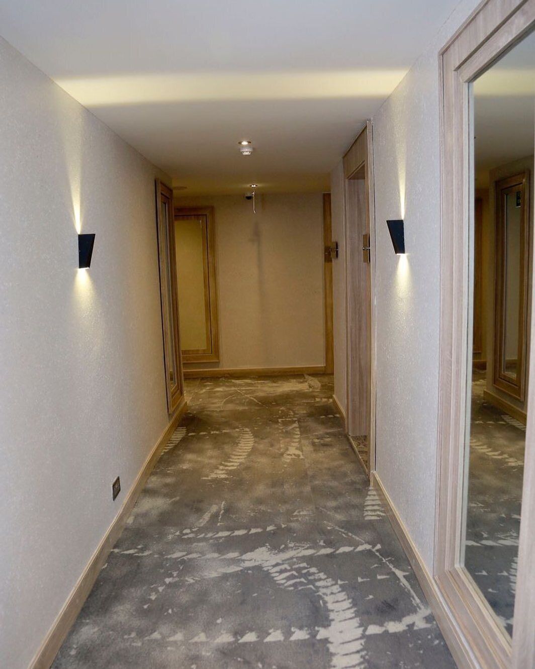 A narrow hallway with light-colored walls and a patterned carpet. There are two wall-mounted light fixtures on either side.
