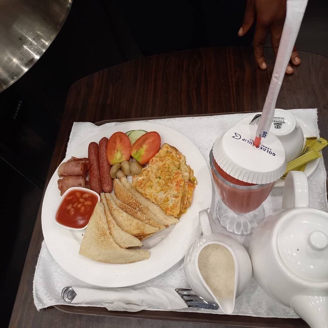 Un plato de comida que incluye huevos, salchichas, tomates y frijoles, una bebida con pajita y una tetera sobre una mesa.