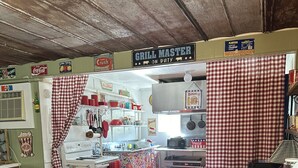 Una zona de cocina con un cartel que dice “GRILL MASTER ON DUTY” y otro que indica “ORDENAR AQUÍ”. Hay dos taburetes frente al mostrador.