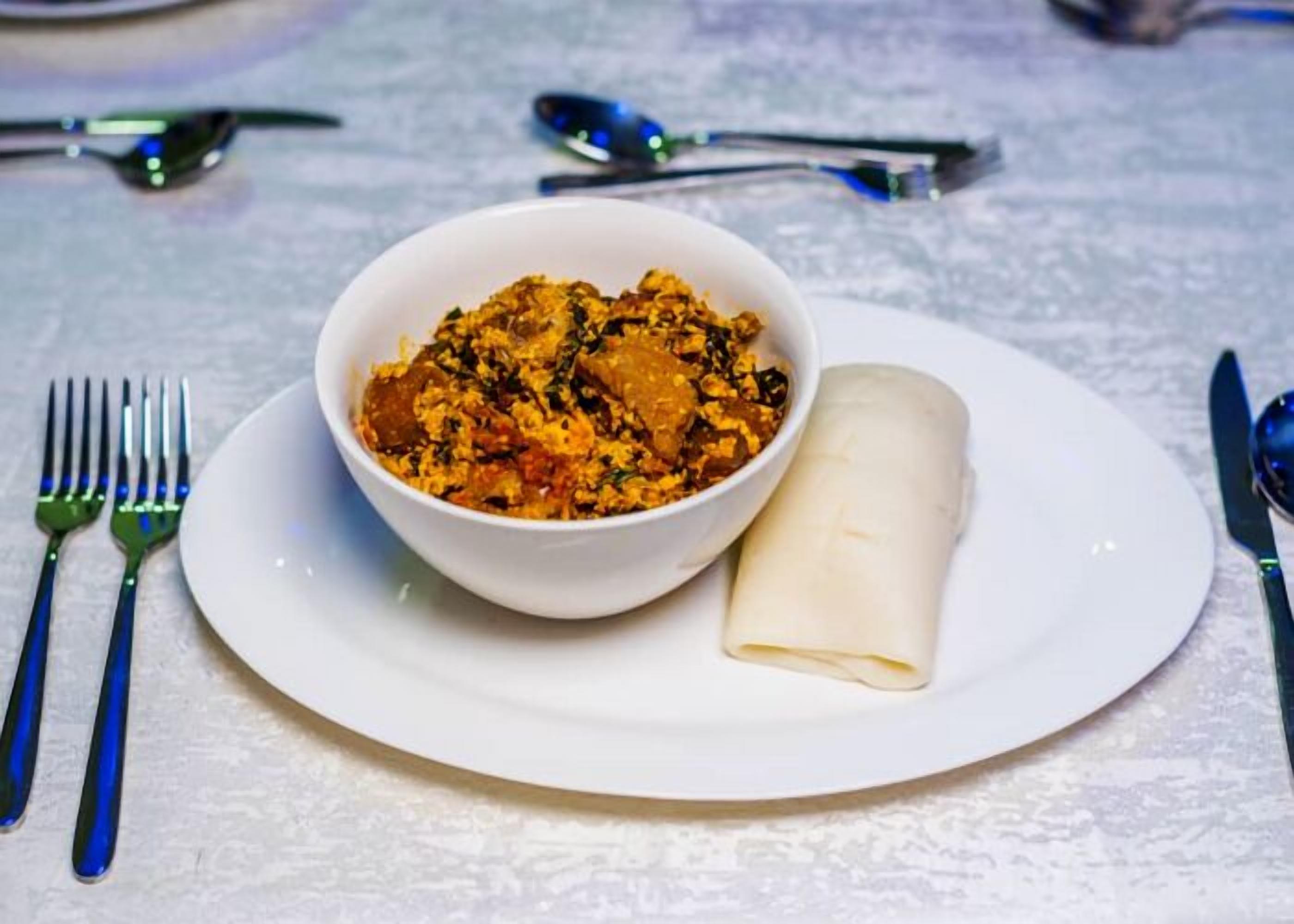 A white plate with a bowl of food and a rolled item, possibly a pastry, on a table with a white tablecloth.