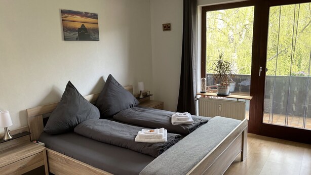 Un dormitorio con una cama de madera, ropa de cama gris, una mesita de noche con lámpara, una pequeña mesa con lámpara y vistas a la vegetación a través de una puerta corrediza.