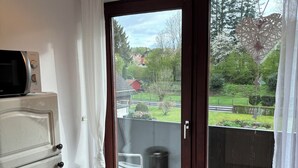 Una habitación con suelo de madera, una puerta corredera de vidrio a un balcón, un radiador blanco y un cubo de basura pequeño.
