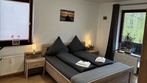Un dormitorio con cama, mesitas de noche, lámpara, un cuadro en la pared y una ventana con cortinas.