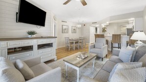Een woonkamer met een flatscreen televisie, een plafondventilator, een eethoek met een tafel en stoelen, en een keuken met een koelkast en een lamp.