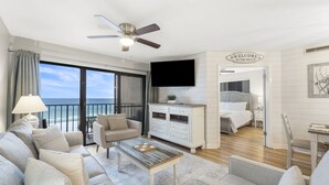 Een ruime woonkamer met een groot raam met uitzicht op het strand, een plafondventilator, een flatscreen-tv en een gezellige zithoek.