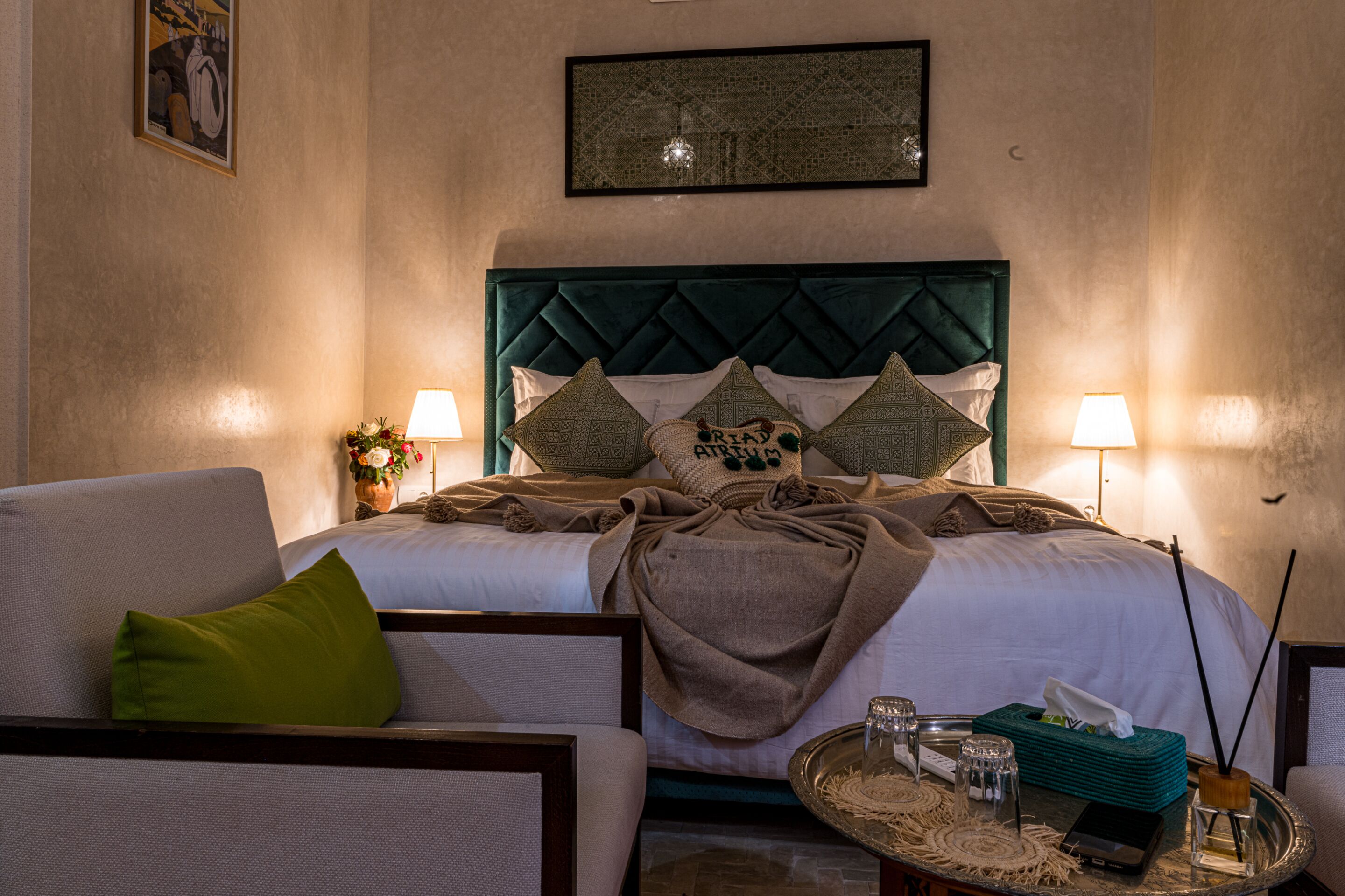 Habitació d'hotel amb un llit, dues làmpades de nit, una manta verda, una taula de nit amb una bossa de mocadors, gots i un objecte decoratiu.