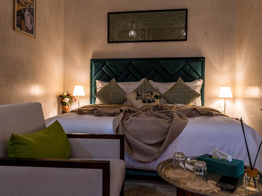 Habitación de hotel con una cama, dos lámparas de noche, un edredón verde, una mesita de noche con un box de pañuelos, vasos y un objeto decorativo.