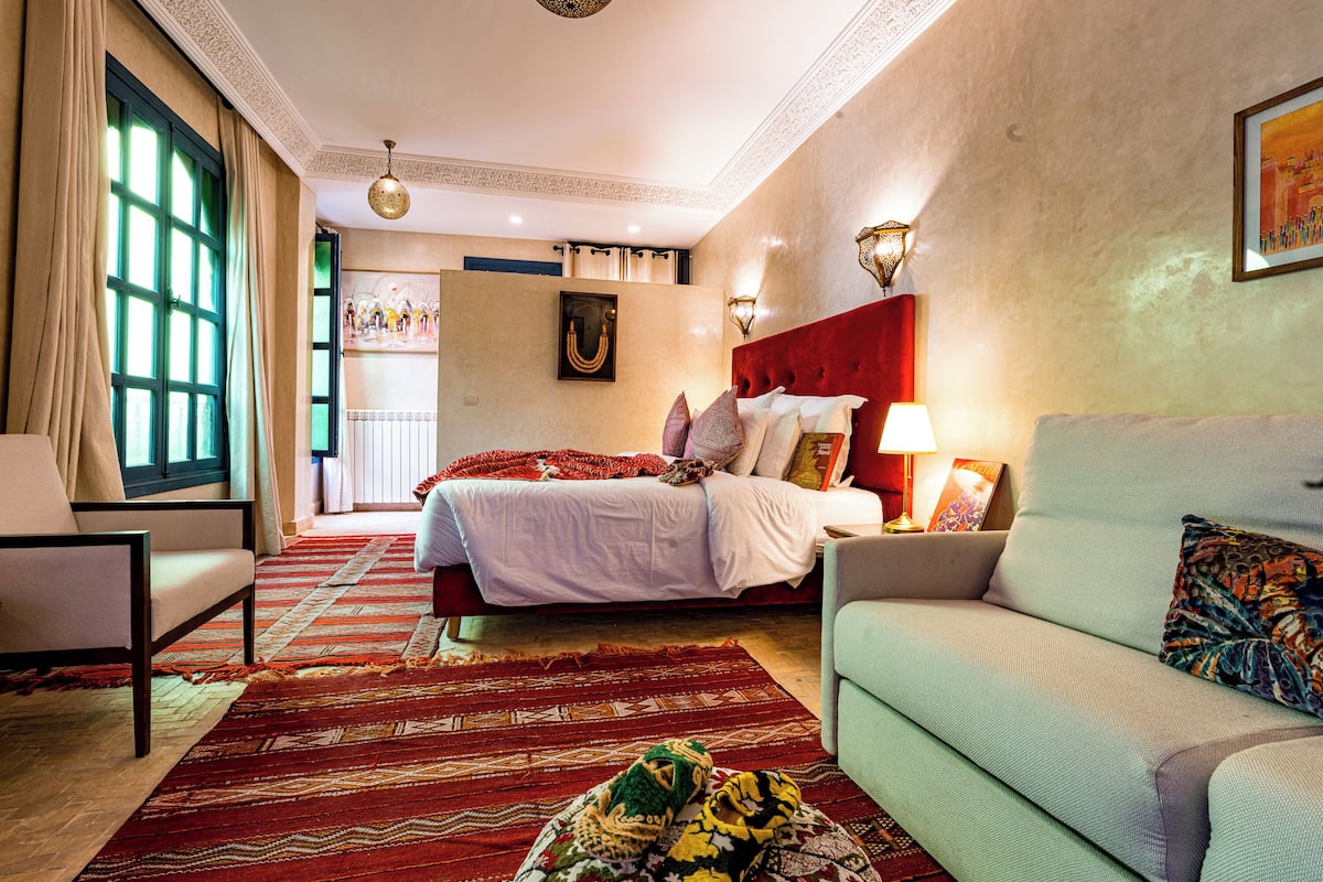 A hotel room with a large bed, a sofa, a patterned rug, and a view of the city through the window.