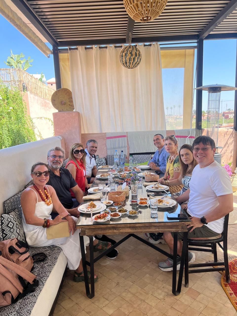 Un grupo de personas sentadas alrededor de una mesa con diversos platos, bajo un porche cubierto con vistas al exterior.