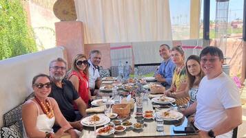 Un grupo de personas sentadas alrededor de una mesa con diversos platos, bajo un porche cubierto con vistas al exterior.