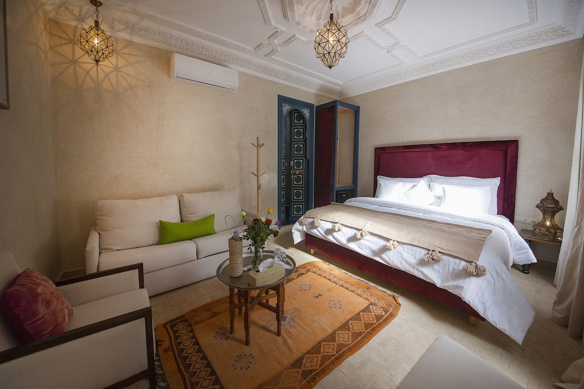 A bedroom with a bed, a sofa, a small table with a vase, and a patterned rug.