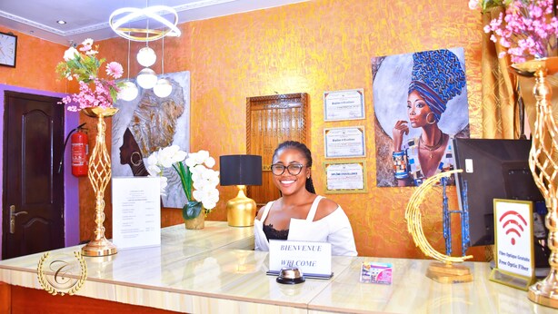 A reception desk with a 'Bienvenue' sign, a gold lamp, a framed certificate, and a 'Free Optic Fiber' sign. - CONSTELLATION HOTEL (Dschang)