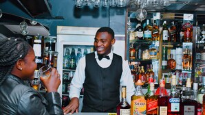 A bar with a bartender and a customer. The bar has shelves stocked with various bottles of alcohol. - CONSTELLATION HOTEL (Dschang)