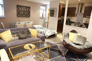 A modern living room with a sofa, armchair, and coffee table, a dining area with a table and chairs, and a kitchen area with cabinets and a stove. - Apartment Sleeps 4 With Walk-in Shower (Birmingham)