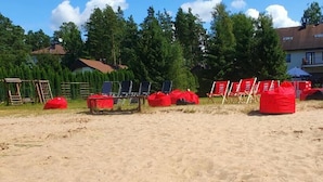 A sandy beach area with red chairs and umbrellas, a building with multiple windows and balconies, and a forested background. - Laguna House (Garkalnes novads)