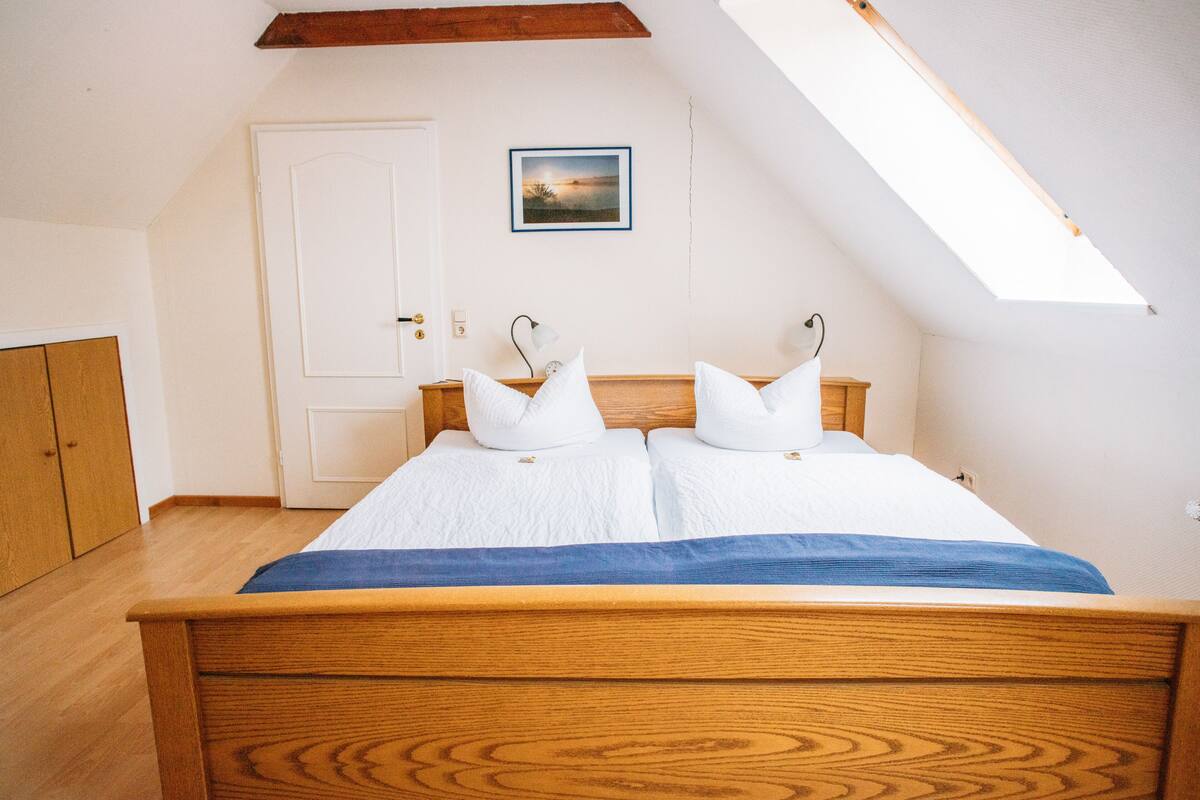 A bedroom with a wooden bed, white bedding, a skylight, a door, and a framed picture on the wall.