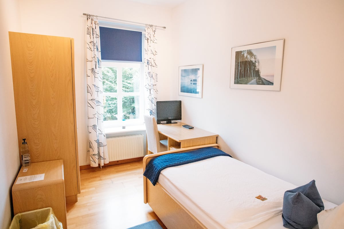 A bedroom with a bed, a desk with a computer, a window with curtains, a radiator, and a door.