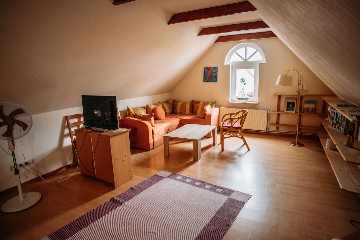 A cozy attic room with a sloped ceiling, a wooden floor, a sofa, a coffee table, a fan, and a window with a view.