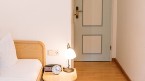 Une chambre d’hôtel avec un lit en bois, une table de chevet avec une lampe, une horloge et une porte visible au fond.