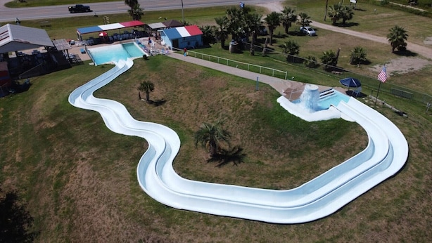 Luchtbeeld van een waterpark met een kronkelende waterglijbaan, een zwembad en omliggende grasvelden met palmbomen.
