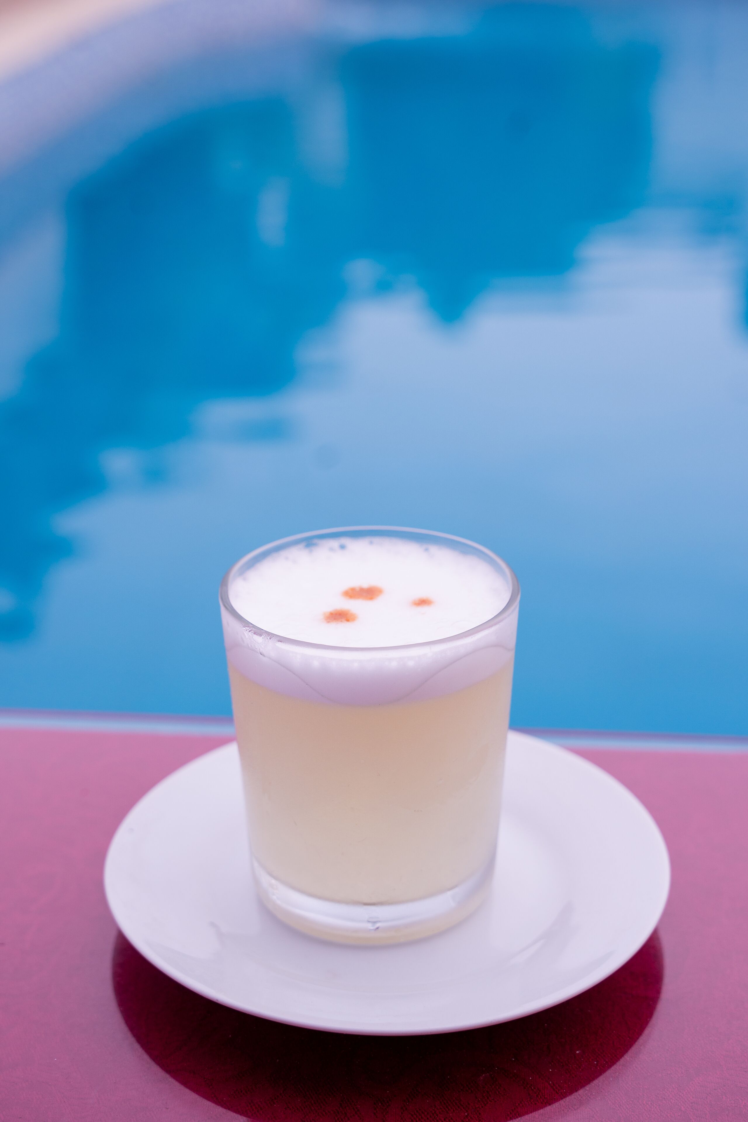 A glass of white beverage on a white saucer by a pool.