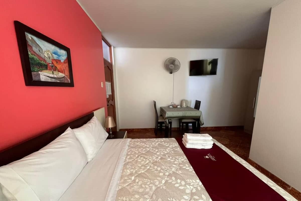 A hotel room with a bed, a dining table with chairs, a TV, and a wall-mounted fan.