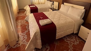 Quarto de hotel com duas camas, uma mesinha de cabeceira, uma luminária e uma janela com cortinas.