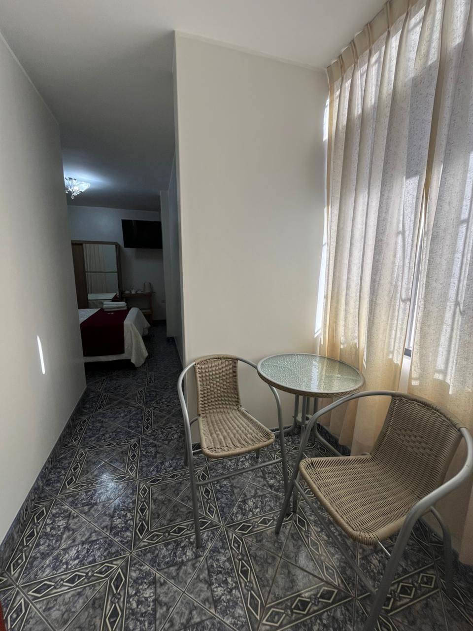 A small, compact room with a bed, a TV, a table with chairs, and a window with curtains.