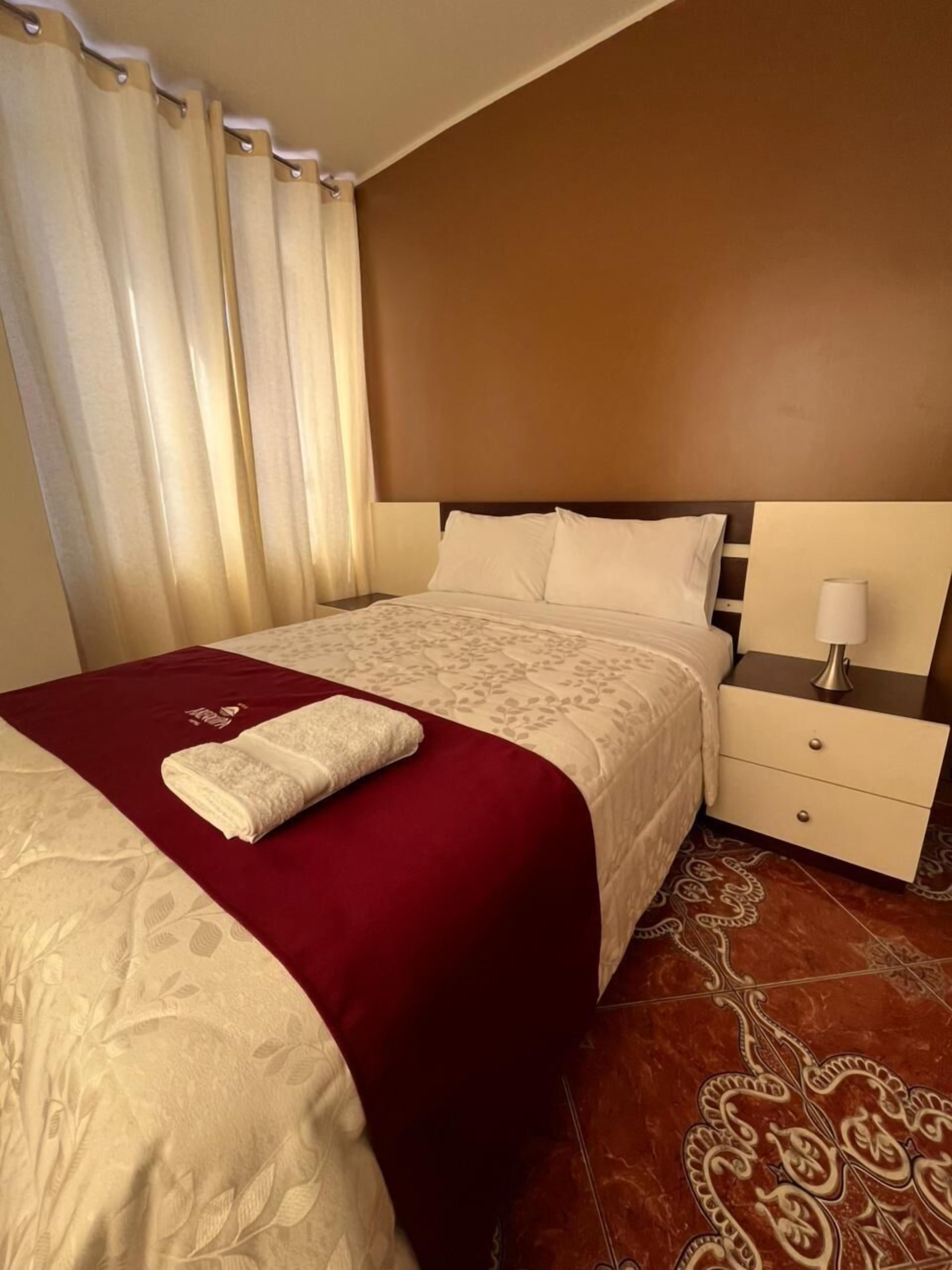 Una cama bien hecha con una colcha roja y blanca, una almohada blanca y una toalla blanca doblada sobre la cama. Hay una mesita de noche con una lámpara y un cajón. La habitación tiene una pared marrón y alfombra estampada.